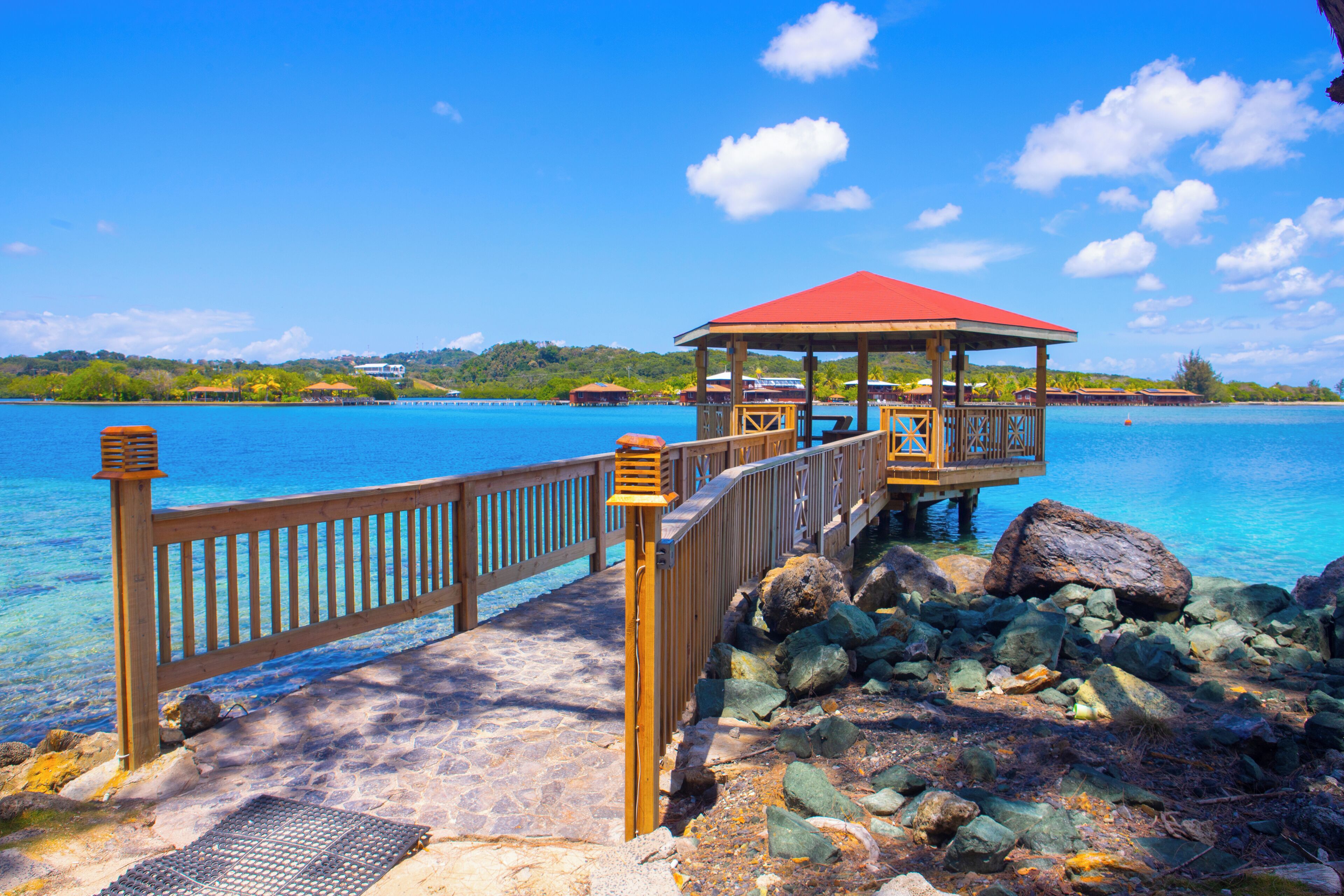 Eine Holzstege führt zu einem Pavillon mit rotem Dach, mit Blick auf ein klares, blaues Meer, in dem Felsen und eine ferne Küste zu sehen sind.