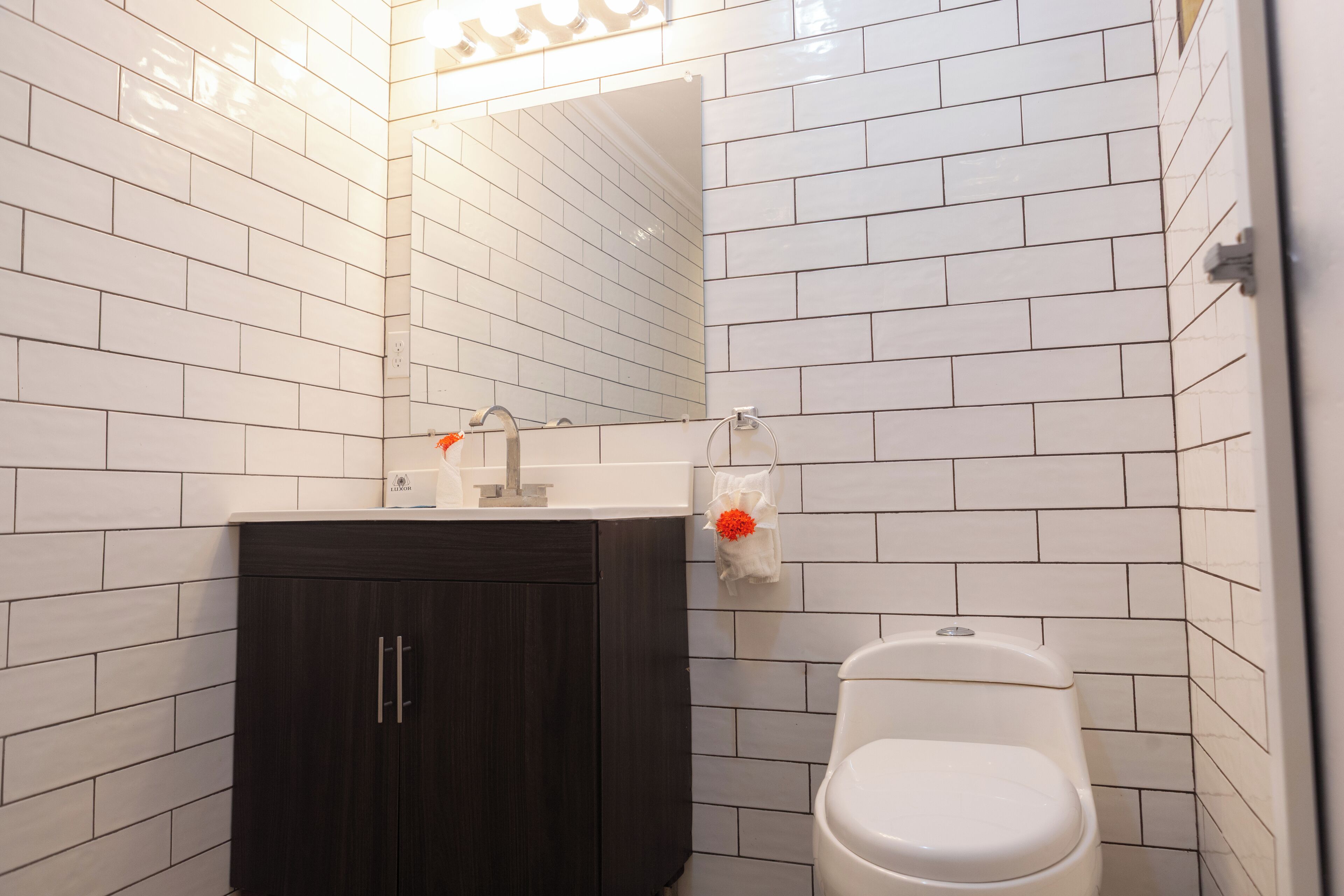 Un baño con paredes de azulejos blancos, un mueble de madera oscura, un lavamanos blanco y un inodoro.