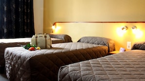 A hotel room with three beds, each with a quilted headboard and a bedside table. There is a suitcase on one bed and a bouquet of flowers on another. The room has a window with curtains and wall-mounted lights. - Hotel Florida (Lourdes)