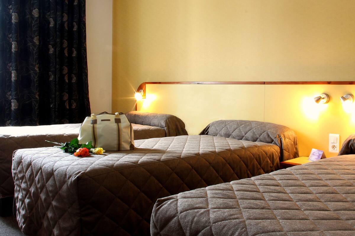 A hotel room with three beds, each with a quilted headboard and a bedside table. There is a suitcase on one bed and a bouquet of flowers on another. The room has a window with curtains and wall-mounted lights.
