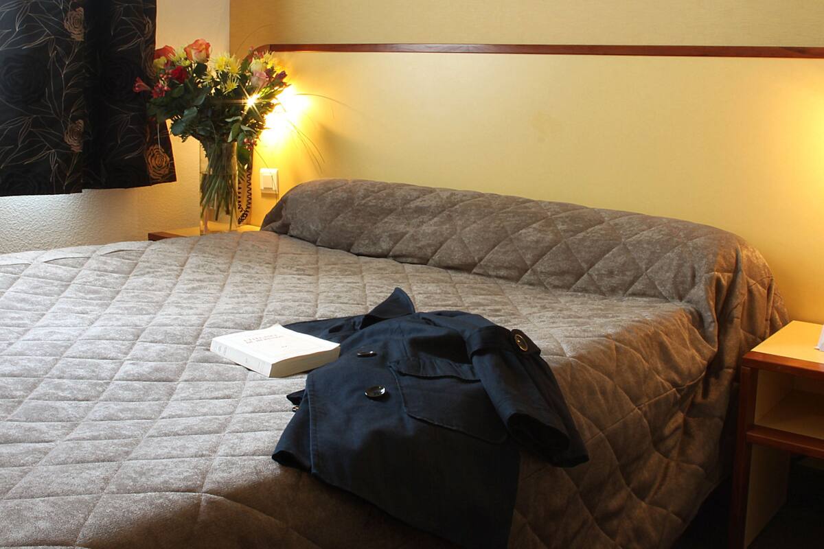A neatly made bed with a quilted cover, a book, and a jacket on it. There is a vase with flowers on the nightstand beside the bed.
