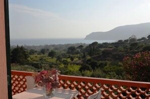 Varanda com mesa e cadeiras, com vista para uma paisagem montanhosa e o mar.