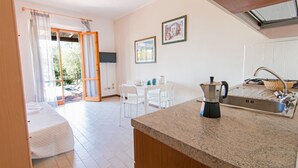 A modern kitchen with a granite countertop, stainless steel sink, and a coffee maker. There is a dining area with white chairs and a table, and a sliding glass door leading to an outdoor area. - Appartamenti Elbamar Lacona (Capoliveri)
