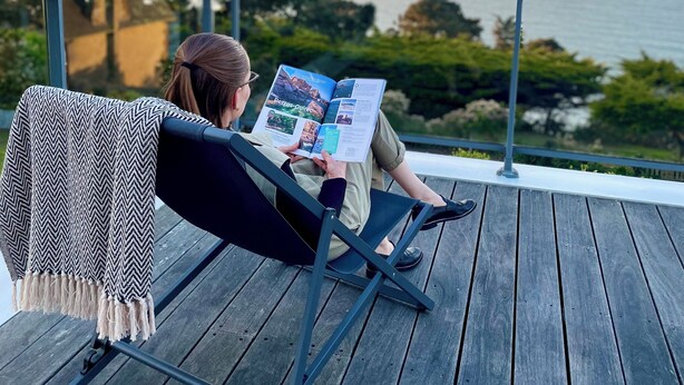 A person reading a magazine on a wooden deck with a railing, overlooking a body of water and trees. - Hôtel Les Costans, The Originals Relais (Perros-Guirec)