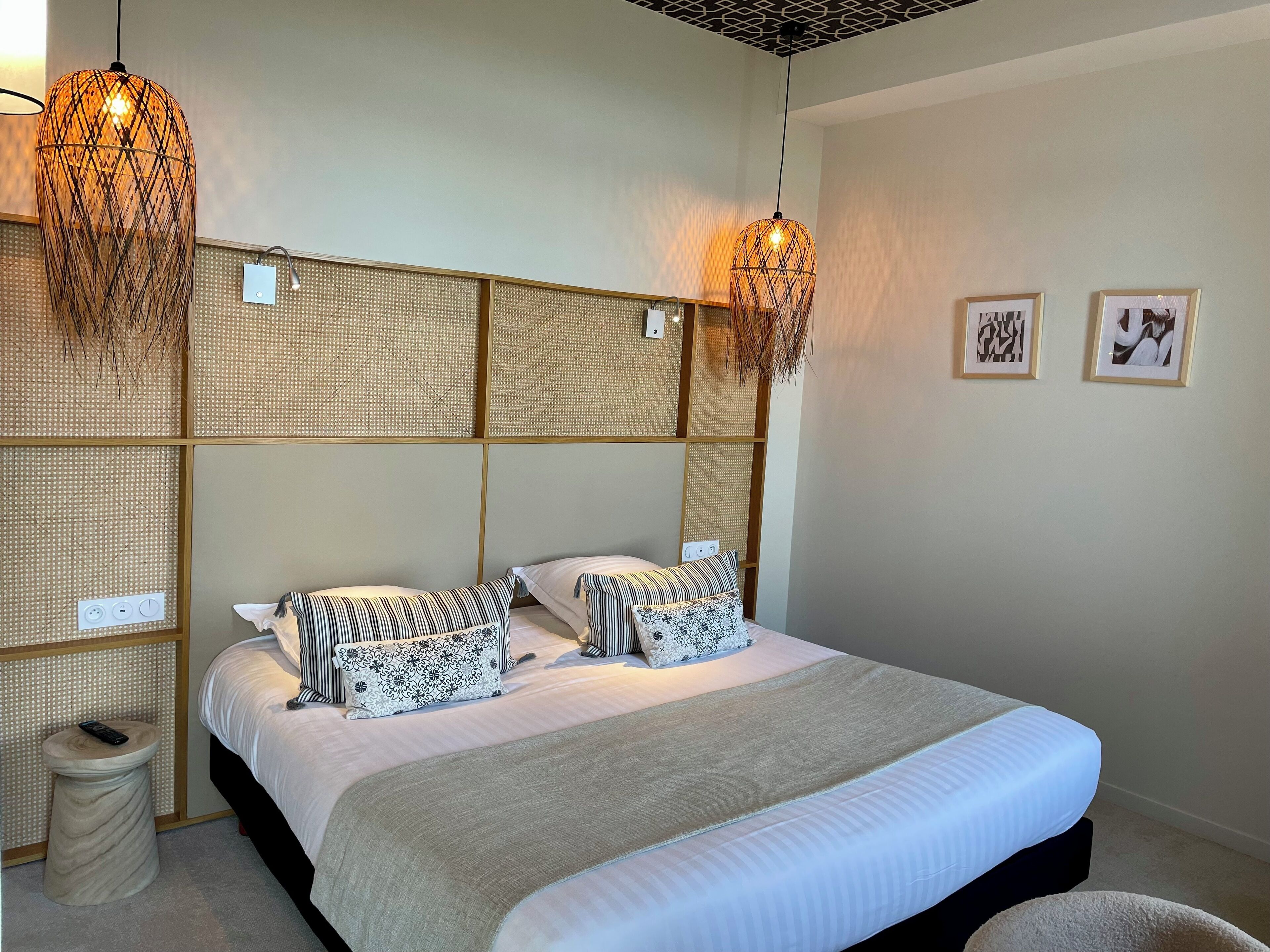 Un lit bien fait, agrémenté d’un coussin à rayures et d’un coussin à motifs floraux, d’une tête de lit en osier et de deux lampes suspendues.