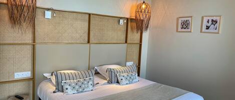 Un lit bien fait, agrémenté d’un coussin à rayures et d’un coussin à motifs floraux, d’une tête de lit en osier et de deux lampes suspendues.