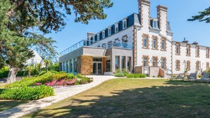 A large, modern building with a glass conservatory, stone facade, and a well-maintained garden with lounge chairs. - Hôtel Les Costans, The Originals Relais (Perros-Guirec)