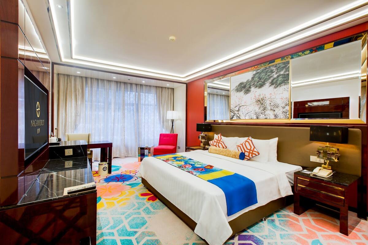 A hotel room with a large bed, a desk with a TV, a red chair, and a colorful carpet.