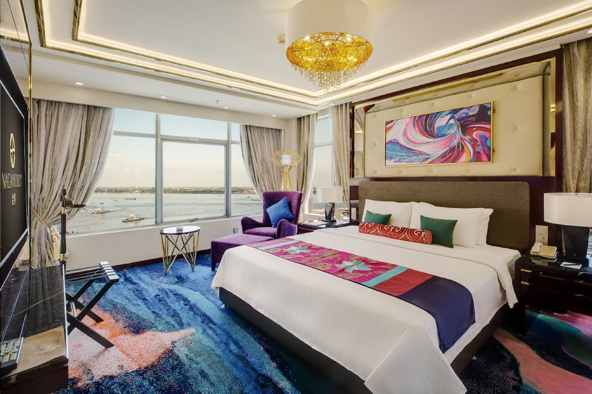 A hotel room with a large bed, a view of the sea, a chandelier, and a painting on the wall.