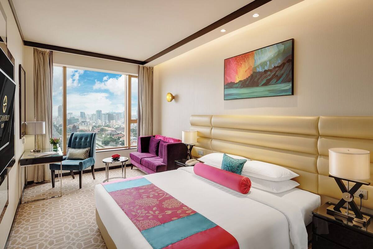 A hotel room with a large bed, a desk, a chair, a sofa, a nightstand, a lamp, and a painting on the wall.