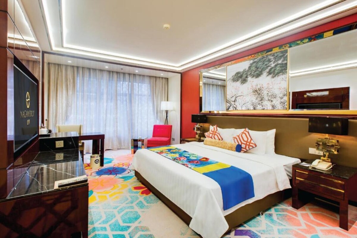 A hotel room with a large bed, a desk with a television, a red chair, and a large mirror.
