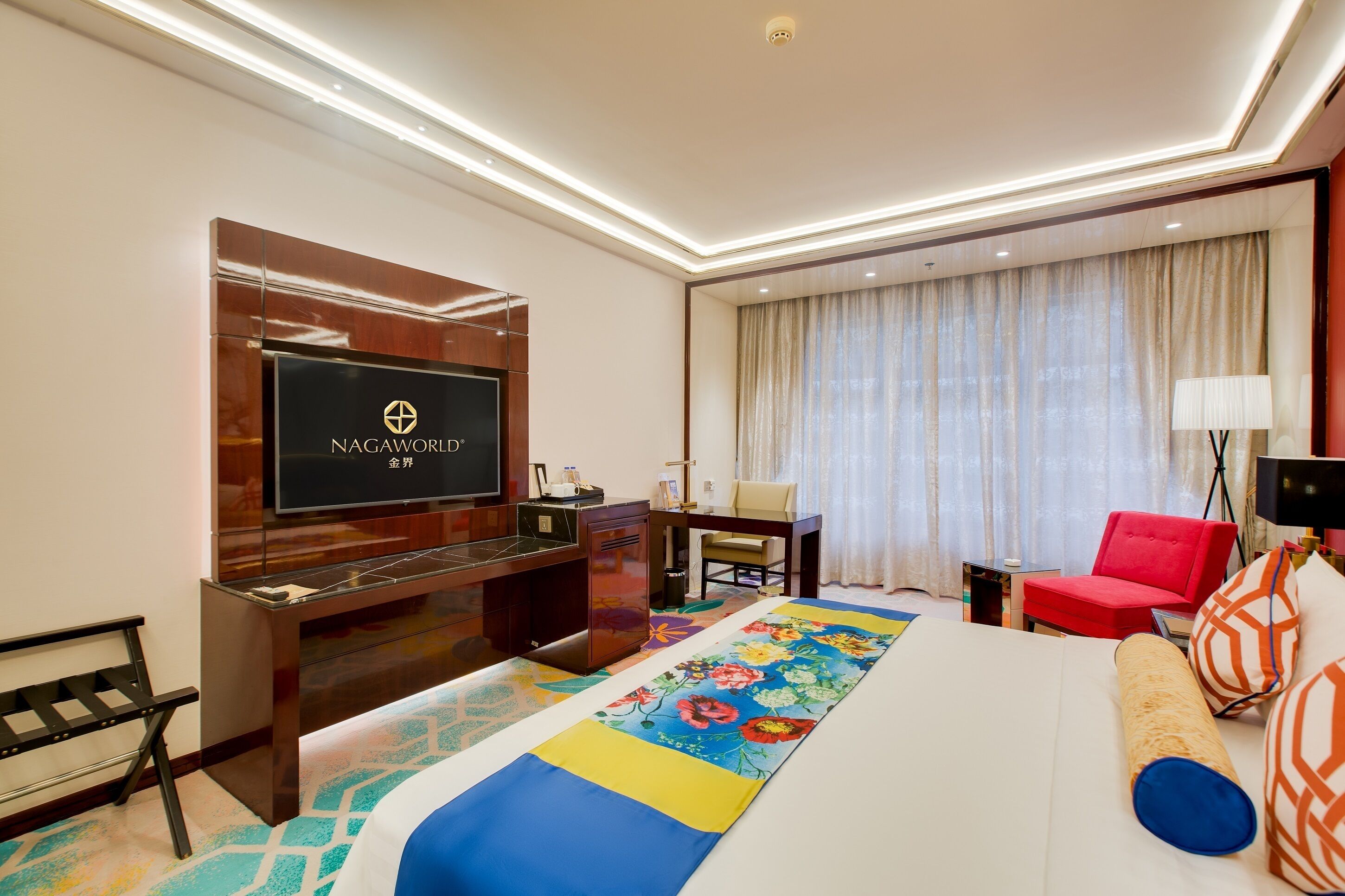 A hotel room with a large bed, a TV mounted on a wooden cabinet, a desk, a red chair, and a colorful area rug.