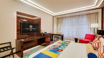 A hotel room with a large bed, a TV mounted on a wooden cabinet, a desk, a red chair, and a colorful area rug.
