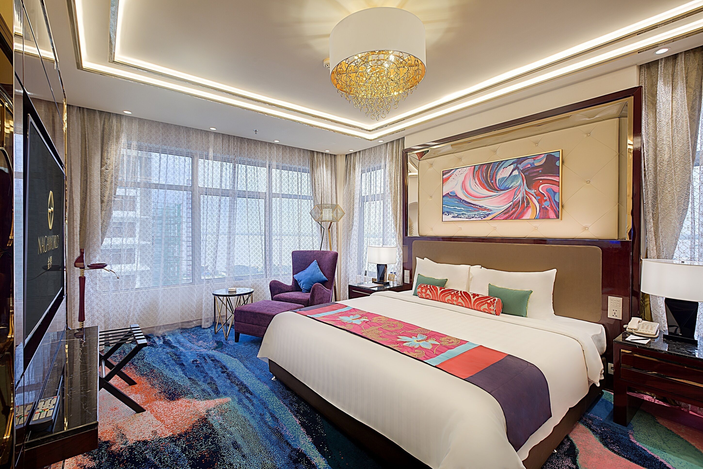 A hotel room with a large bed, a chandelier, a painting, and a view of the city through large windows.