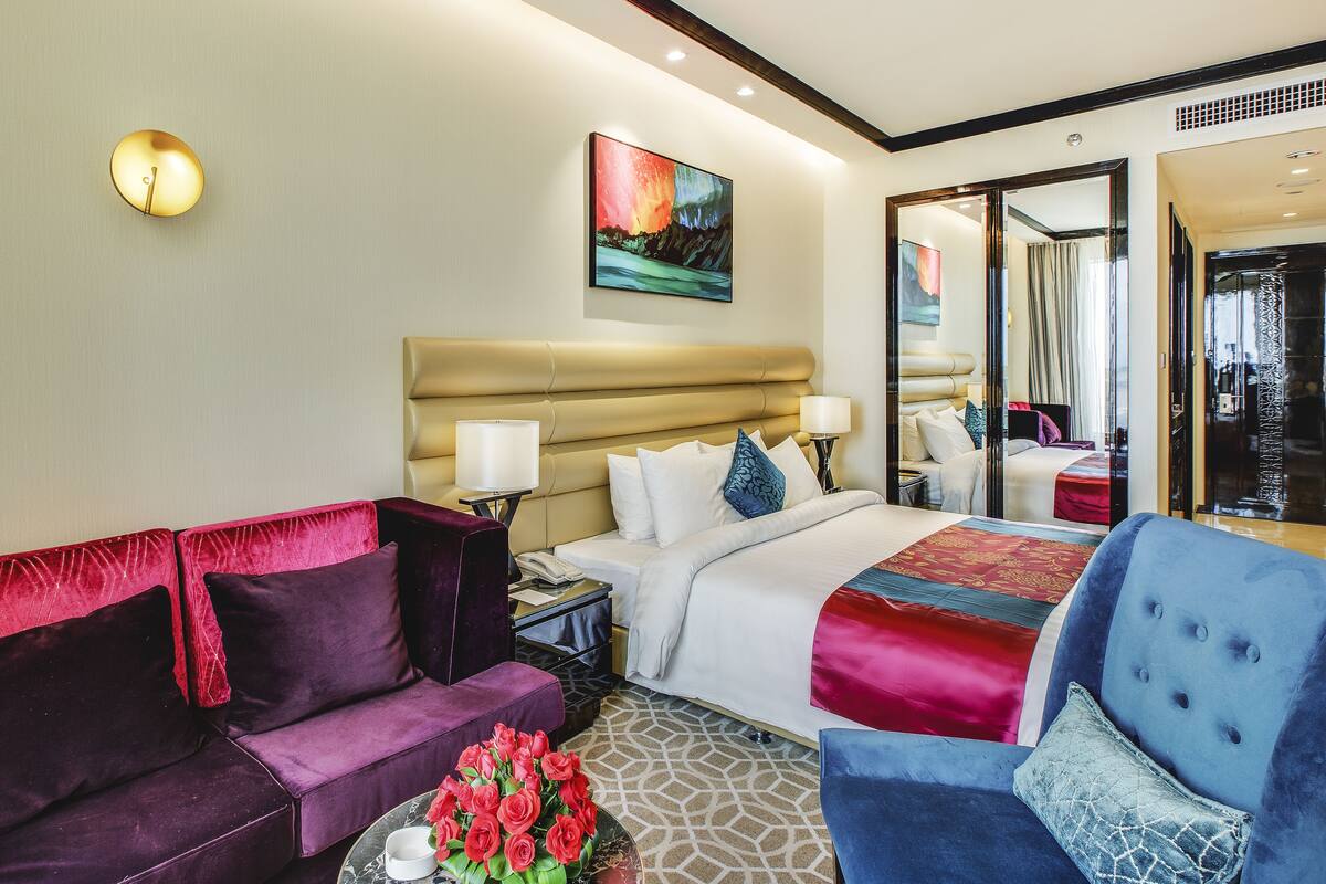 A hotel room with a bed, a sofa, a chair, a nightstand, a lamp, and a painting on the wall.