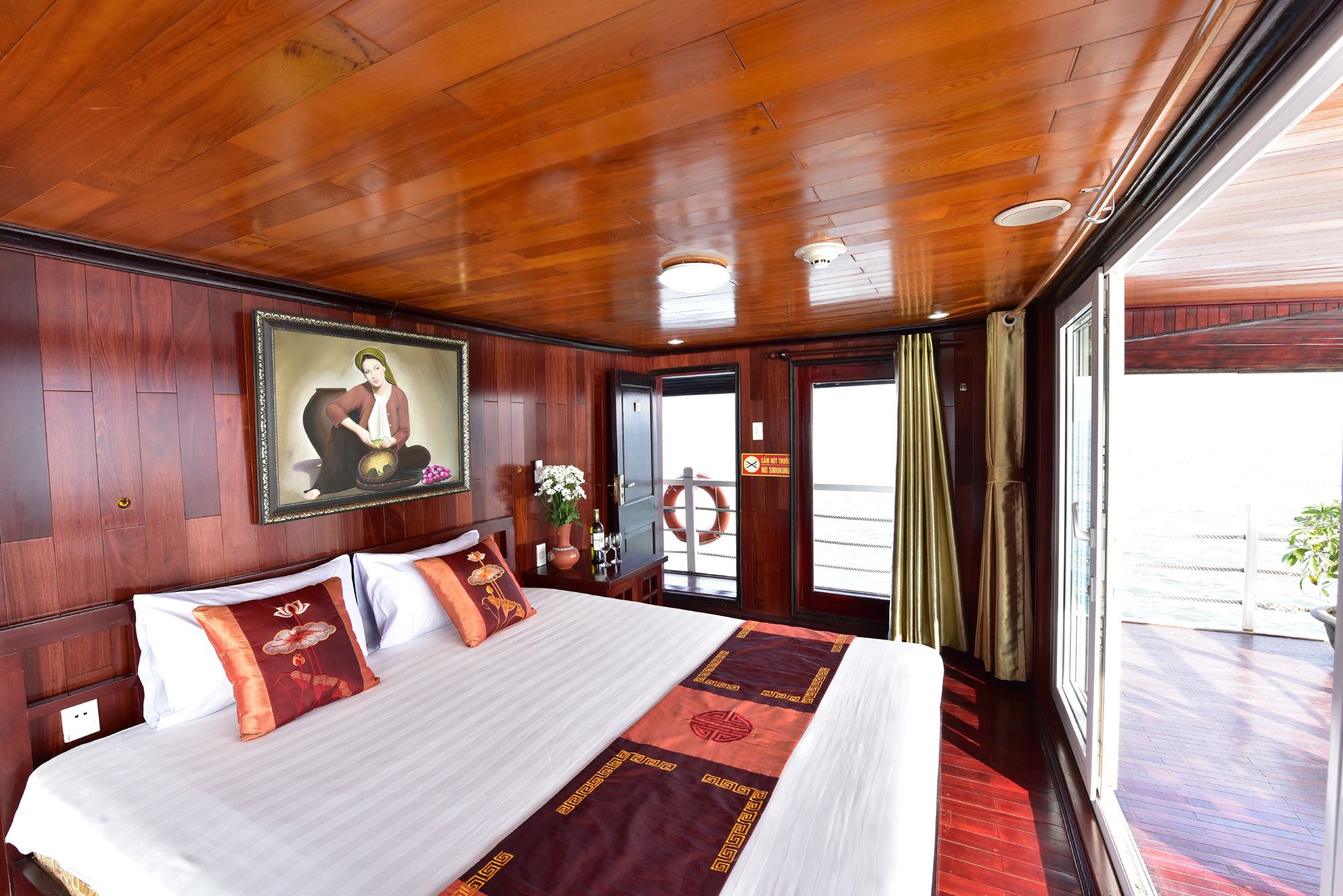 A hotel room with a wooden ceiling, a bed with white and red bedding, a framed picture on the wall, and a view of the outside through a door.