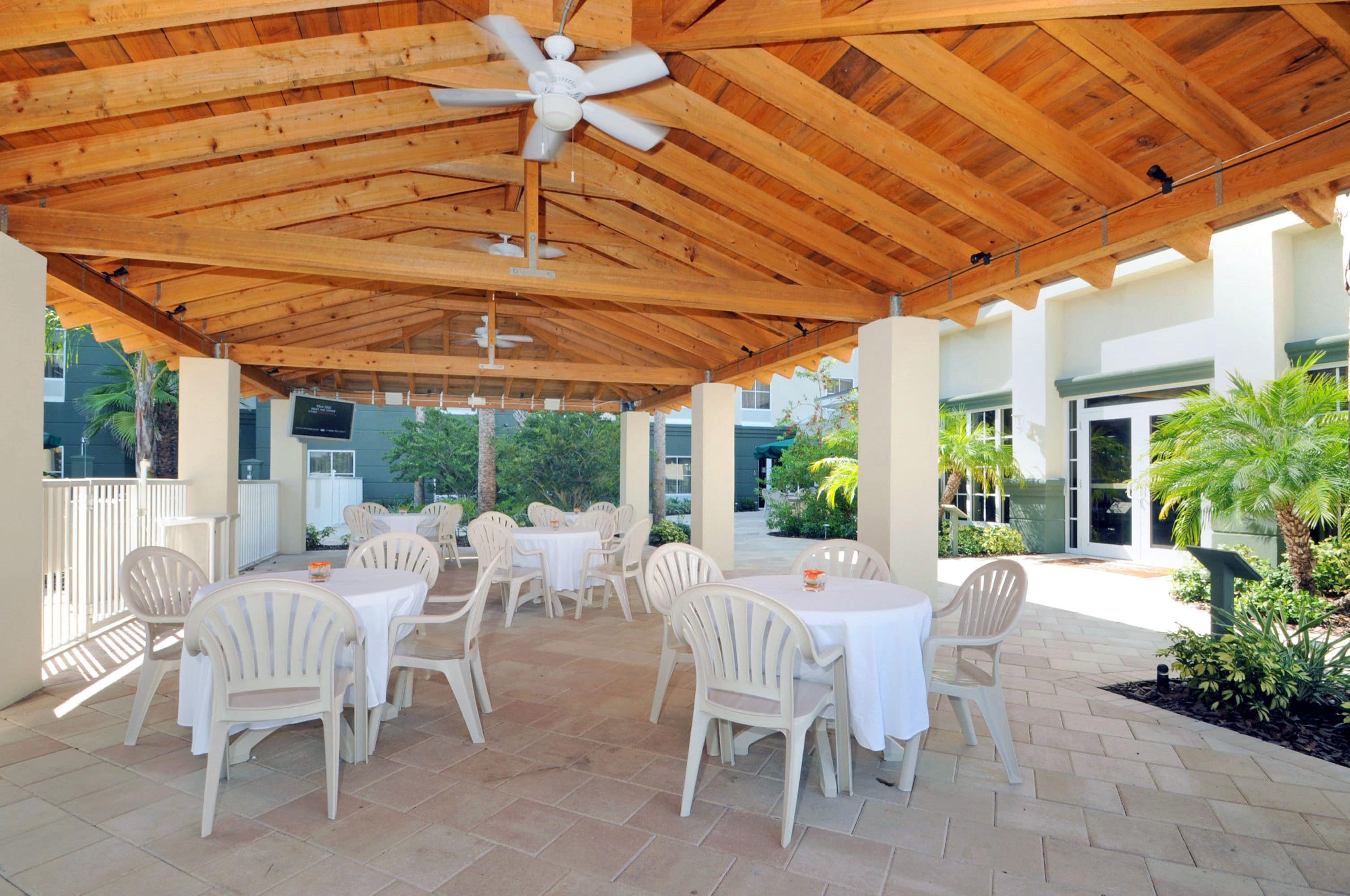 An outdoor patio with wooden beams, white plastic chairs, and tables, surrounded by greenery and a building in the background.