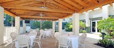 An outdoor patio with wooden beams, white plastic chairs, and tables, surrounded by greenery and a building in the background.