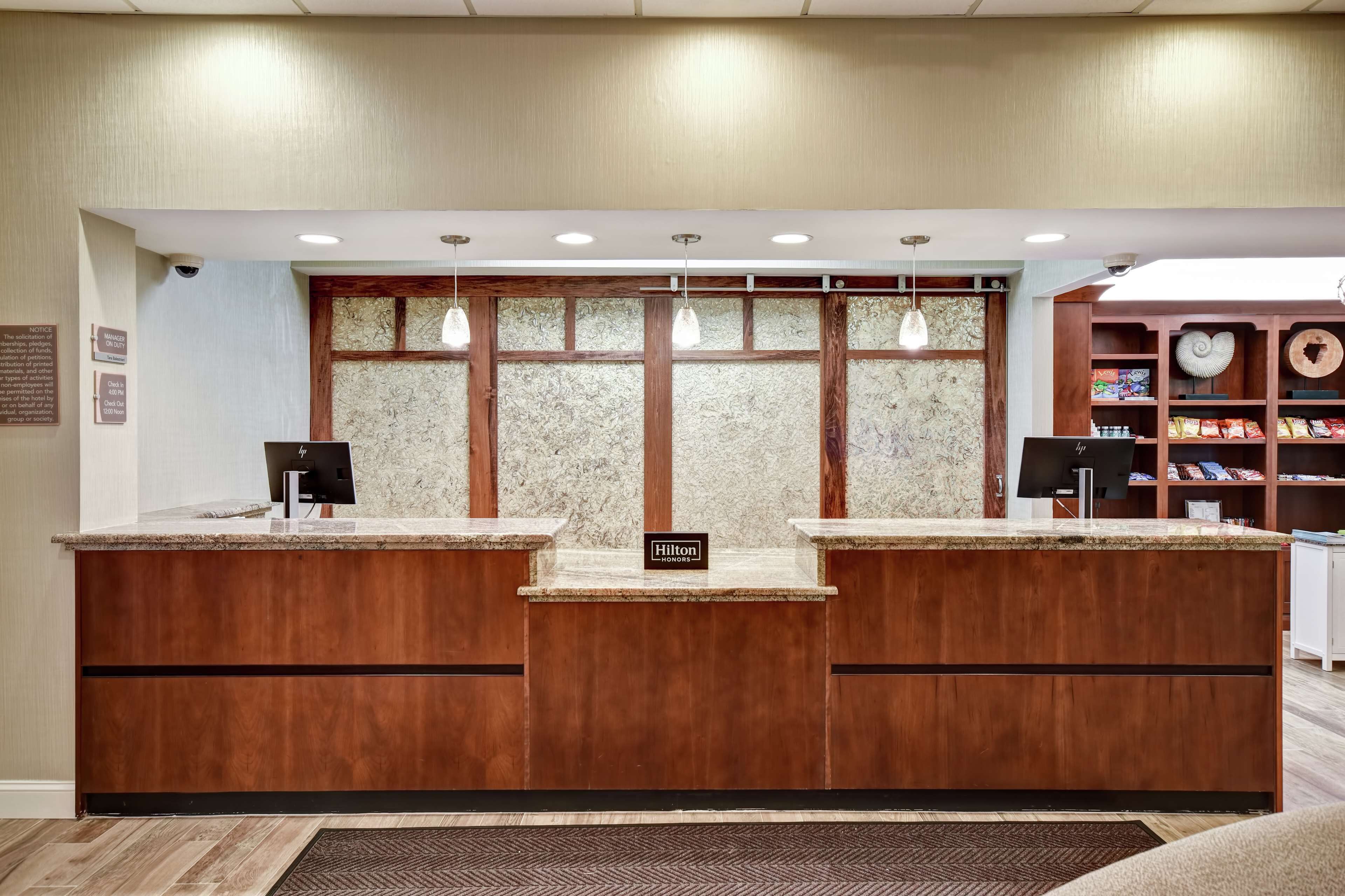 A hotel reception desk with a wooden finish, a marble countertop, and a sign indicating it's a Hilton hotel. There are three pendant lights above the counter, and a security camera is mounted on the wall to the left.