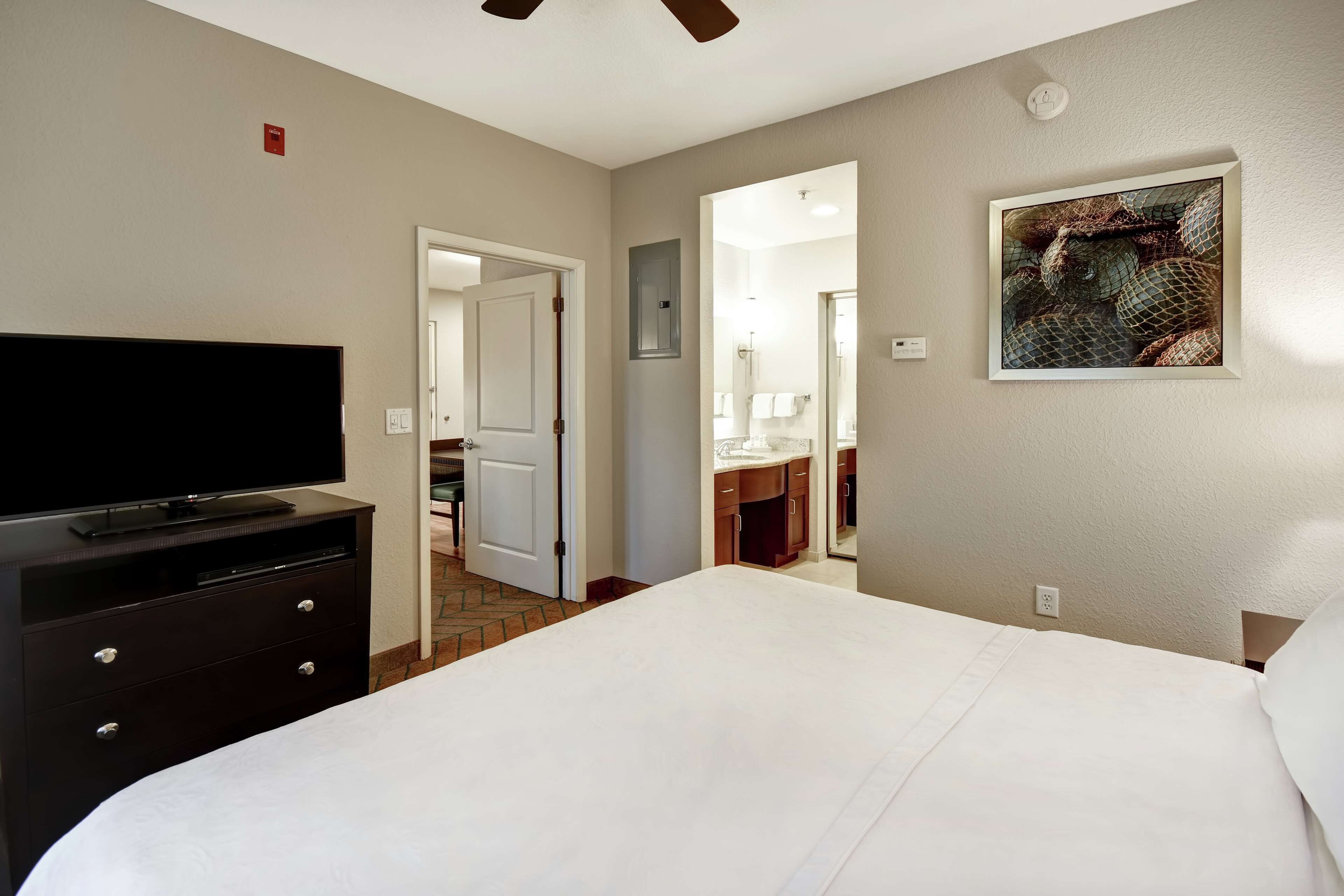 A hotel room with a bed, a dresser, a television, a painting, and a bathroom visible through an open door.