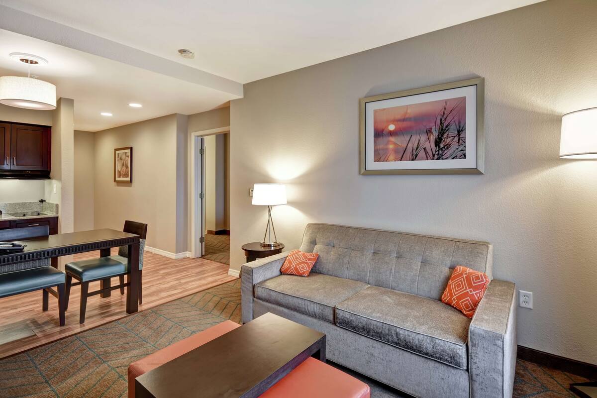 A modern hotel room with a grey sofa, a dining table with chairs, a small side table with a lamp, and a framed picture on the wall.