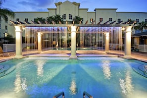 A pool area with a covered seating area, palm trees, and a building in the background.