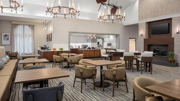 Un hall d’hôtel moderne doté de plusieurs espaces de détente, notamment un long canapé, ainsi que de nombreuses tables et chaises. On aperçoit à l’arrière un coin cuisine, un téléviseur à écran plat fixé au mur et une cheminée.