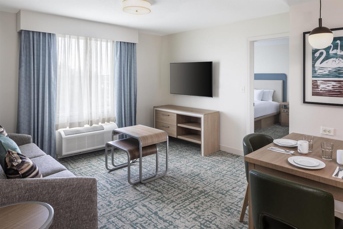 Habitación de hotel con sofá, escritorio, mesa de comedor para dos personas, televisor de pantalla plana y ventana grande con cortinas.