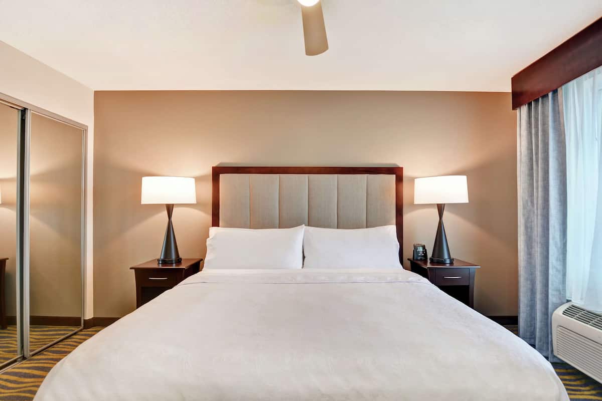 Habitación de hotel con una cama grande, dos lámparas de noche, un ventilador de techo, un espejo y una ventana con cortinas.