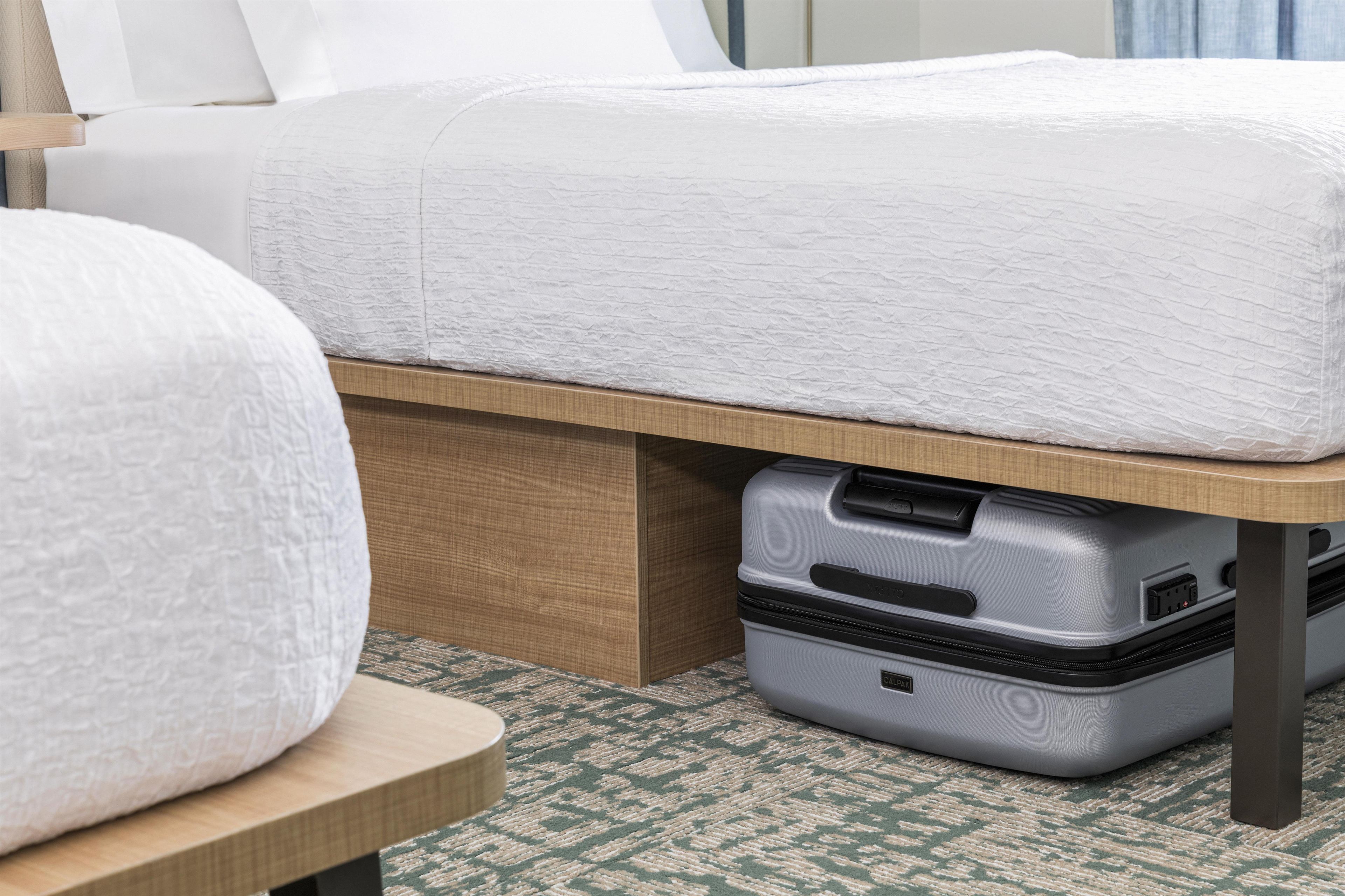 A bed with a white textured bedspread, a wooden bedside table, and a grey suitcase under the bed.