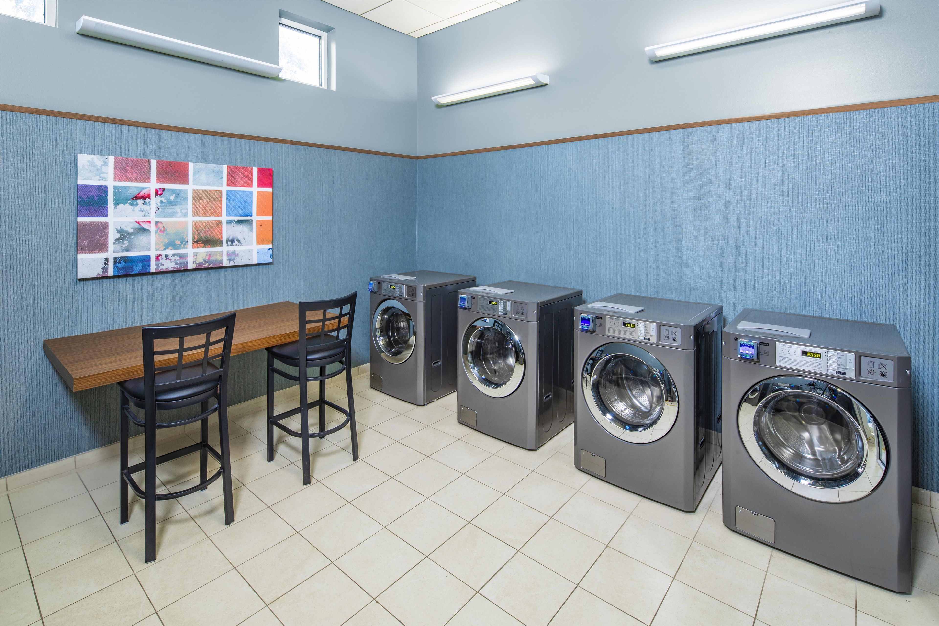 A laundry room with four washing machines, a wooden bench, two chairs, and a wall-mounted artwork.