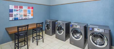 A laundry room with four washing machines, a wooden bench, two chairs, and a wall-mounted artwork.