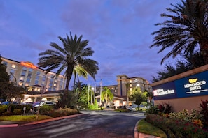 Entrada de hotel com placas da Hilton Garden Inn e Homewood Suites, cercada por palmeiras e paisagismo bem cuidado.