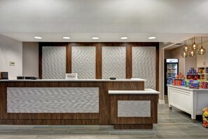 A hotel reception area with a wooden reception desk, a sign indicating Hilton Honors, and a snack bar with various food items.