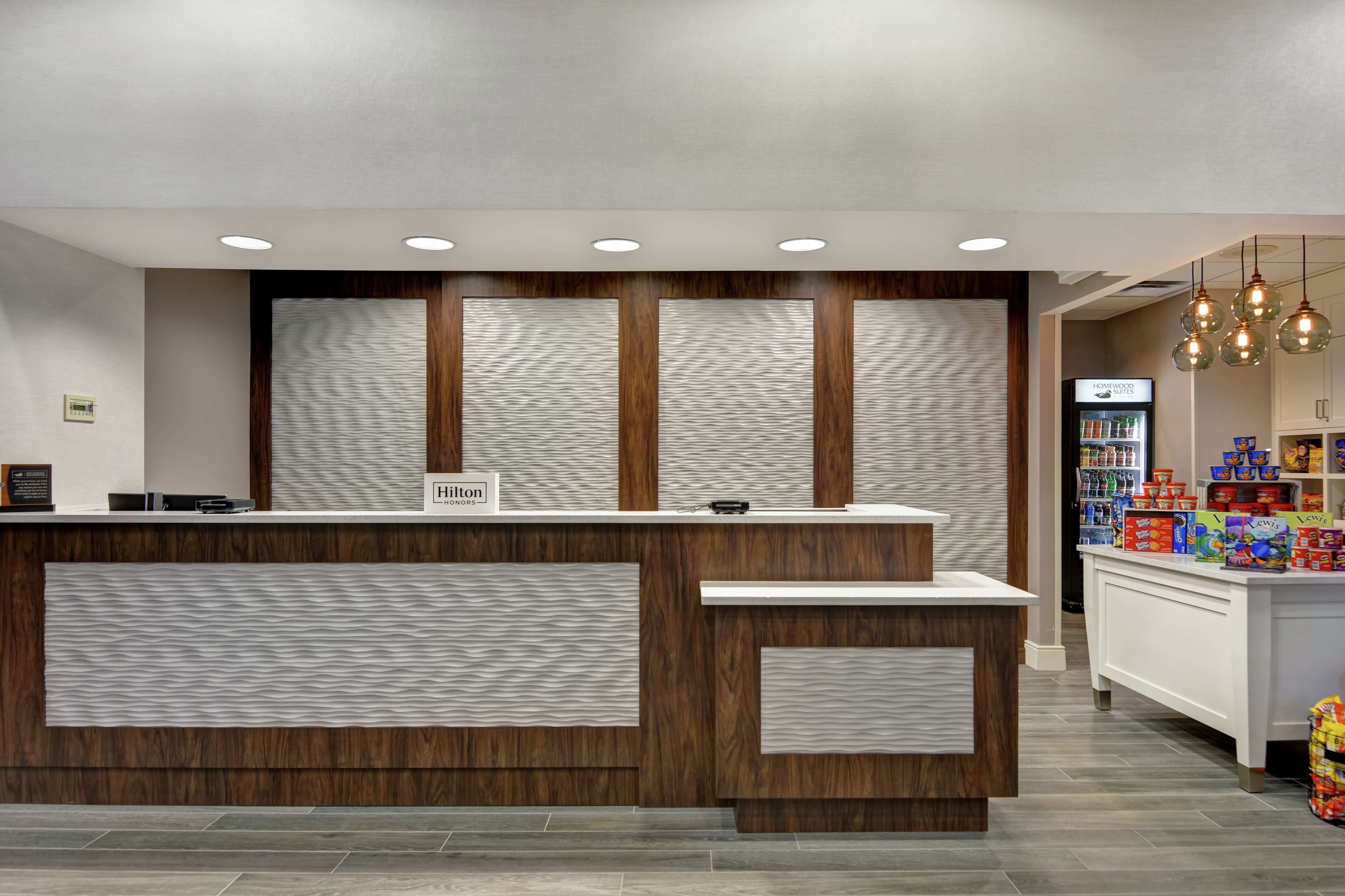 A hotel reception desk with a 'Hilton' sign, a refrigerator stocked with beverages, and a snack bar with various food items.