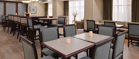 A hotel dining area with multiple tables and chairs, a chandelier, and large windows with curtains.