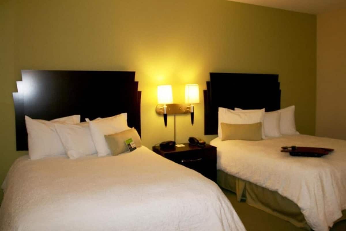 A hotel room with two beds, a nightstand with a lamp, and a wall-mounted light fixture.
