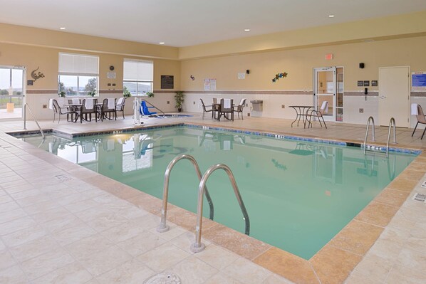An indoor swimming pool with a tiled floor, metal pool ladders, and several tables and chairs around the pool area.