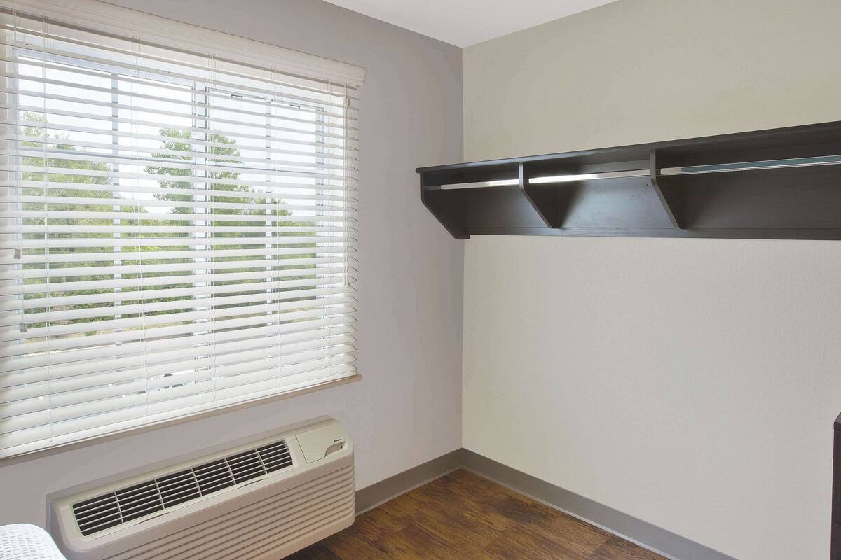 A corner room with a window, built-in shelf, and a wall-mounted air vent.