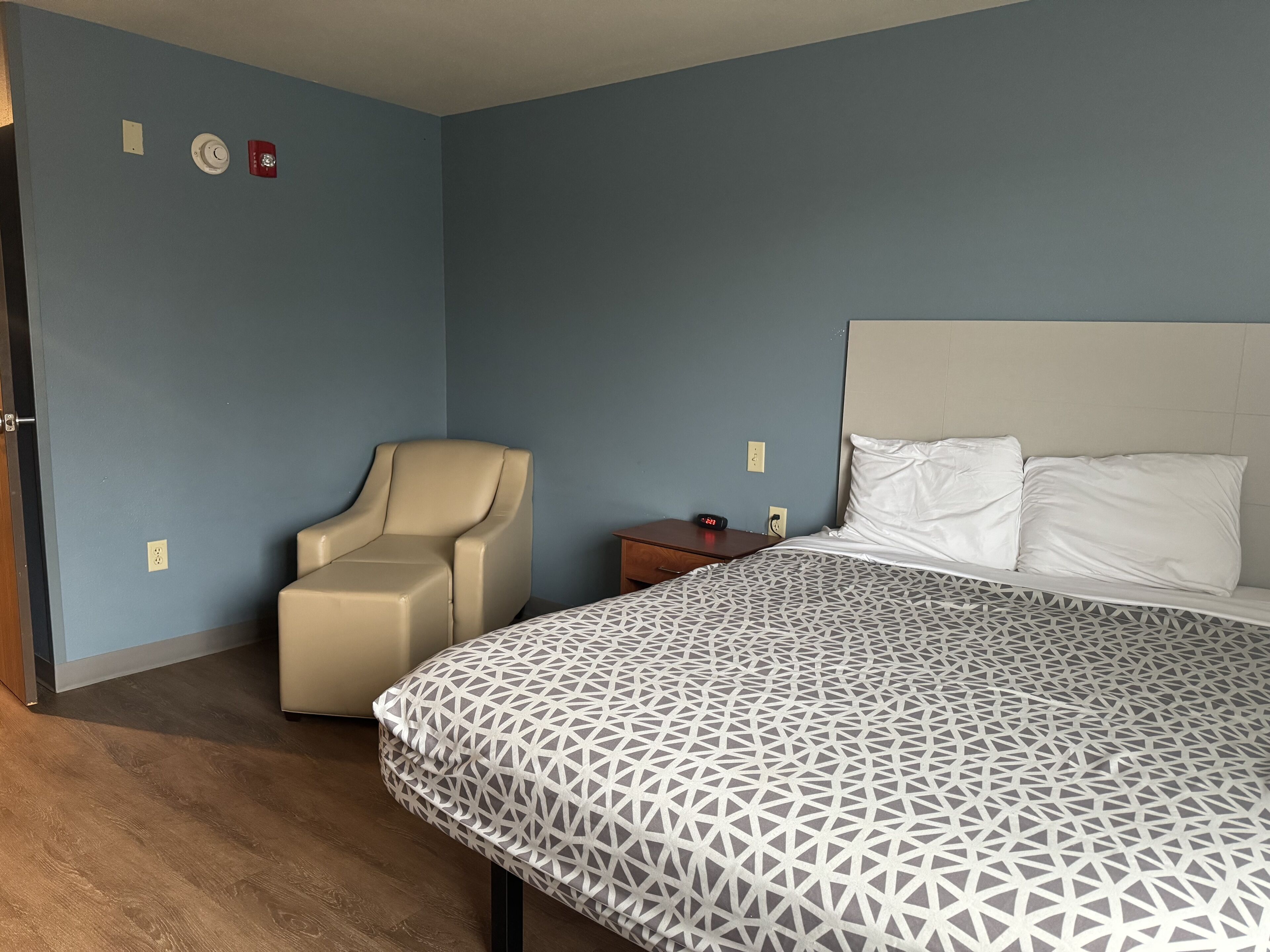A bedroom with a bed, a chair, a nightstand, and a wall with a fire alarm.