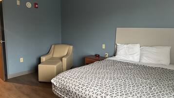 A bedroom with a bed, a chair, a nightstand, and a wall with a fire alarm.