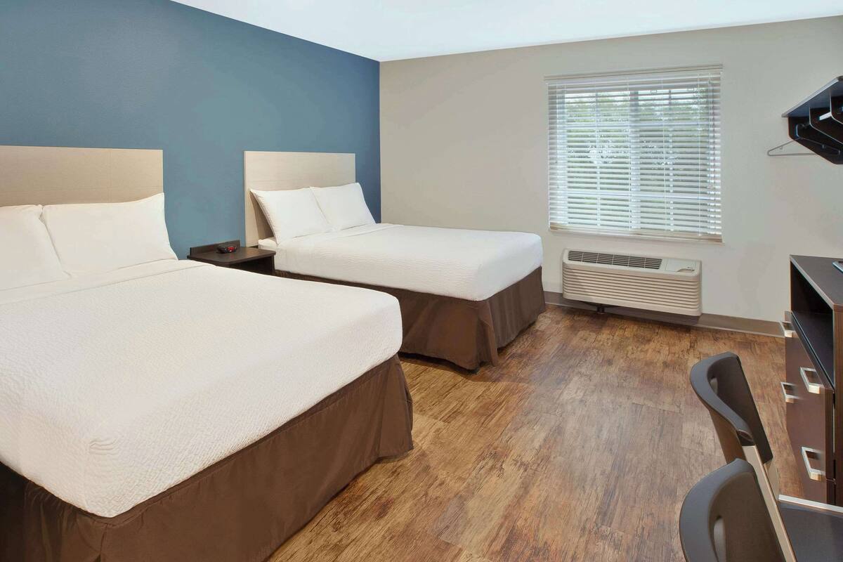 A hotel room with two beds, a desk, and a window with blinds.