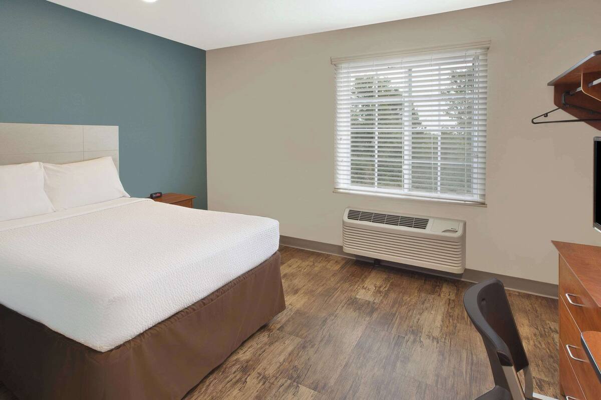 A bedroom with a bed, a desk, a chair, a window with blinds, and an air conditioning unit.