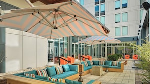 An outdoor seating area with cushioned sofas, striped umbrellas, and a modern building in the background. - Aloft by Marriott Arundel Mills BWI Airport (Hanover)