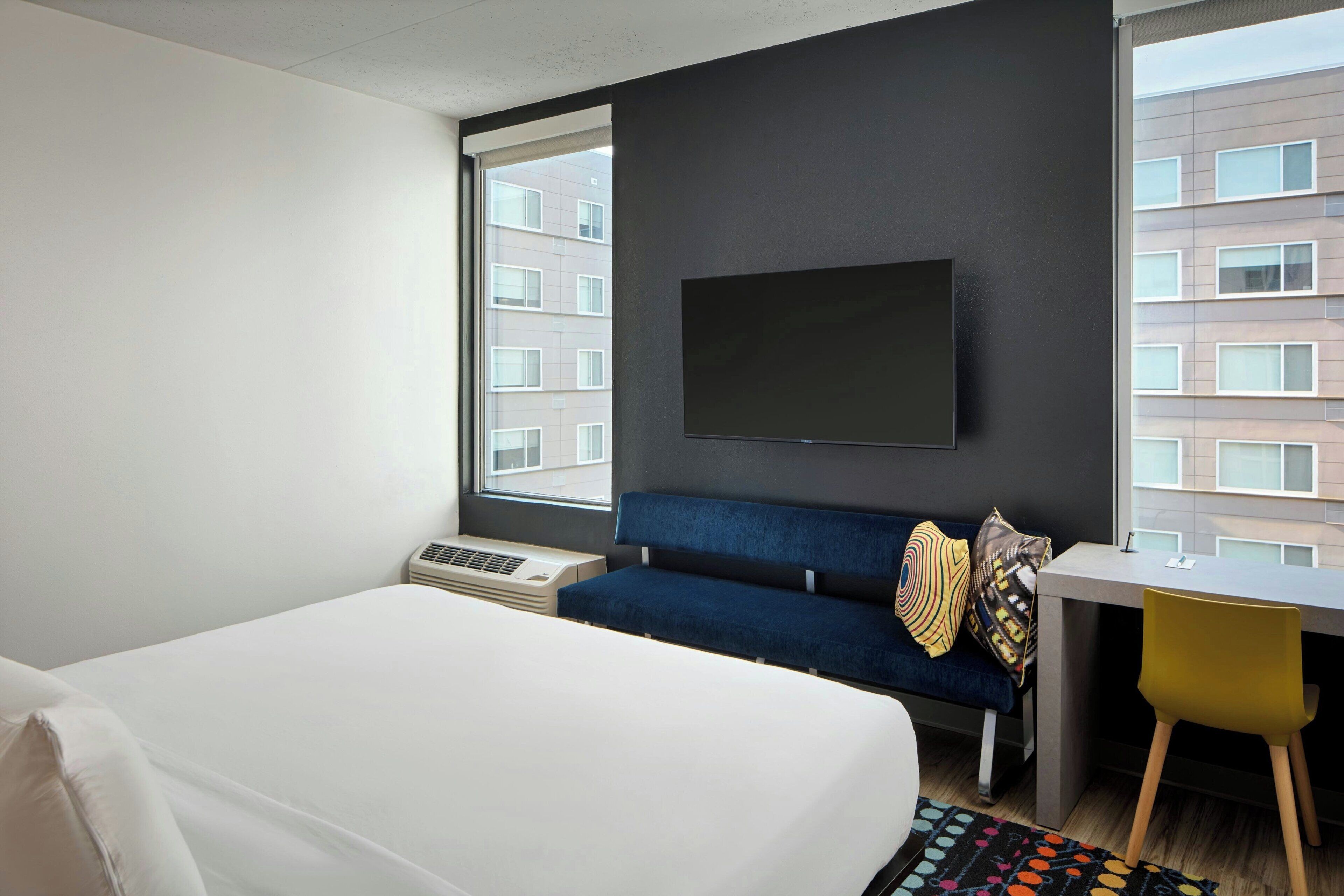 Una habitación de hotel con cama, escritorio, silla, televisor y ventanas amplias.