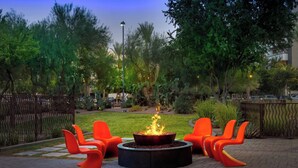 A circular fire pit with orange chairs around it, set in a landscaped garden with trees and a fence. - Aloft by Marriott Tempe (Tempe)