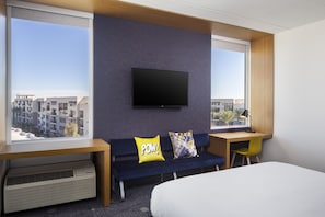 A hotel room with a bed, a desk with a chair, a sofa with two cushions, a television on the wall, and large windows offering a view of buildings. - Aloft by Marriott Tempe (Tempe)
