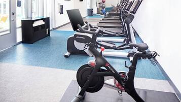 Un gimnasio con varias bicicletas estáticas, piso cubierto de moqueta azul y un amplio ventanal que proporciona luz natural.