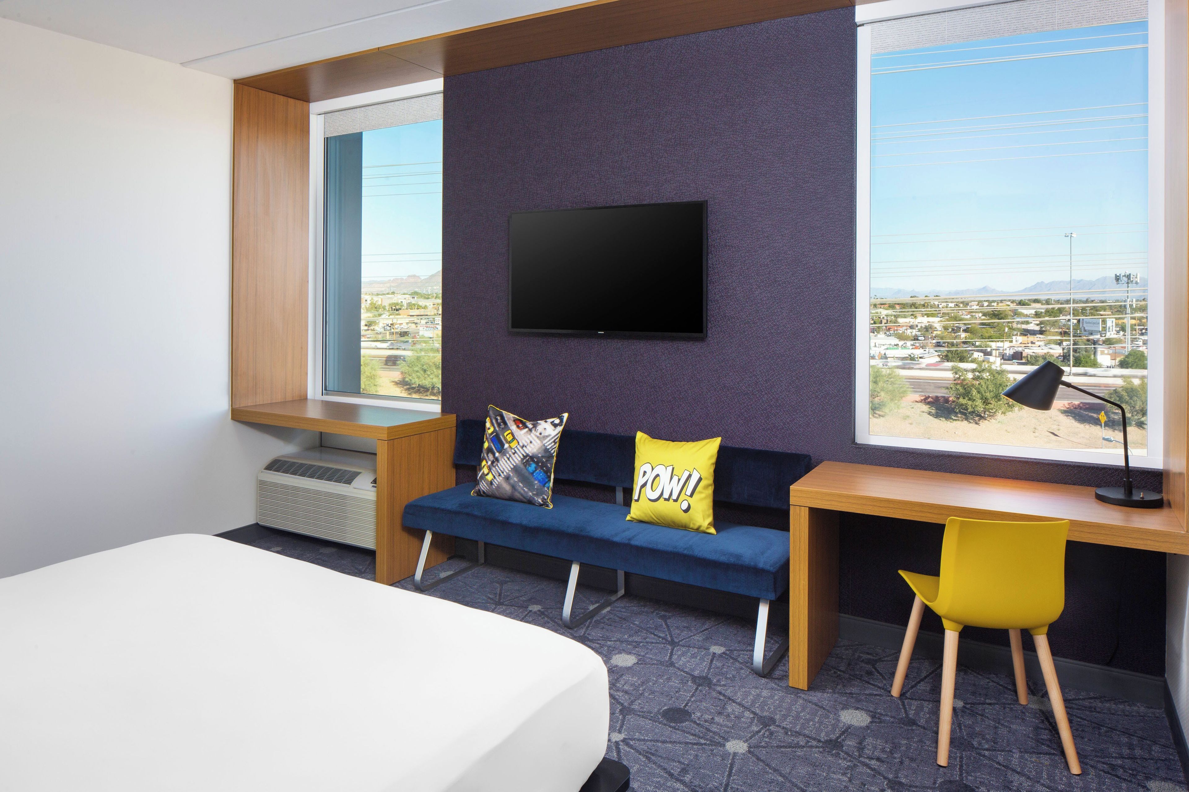 Habitación de hotel con cama, escritorio, silla, sofá con dos almohadas, televisor en la pared y vista panorámica de la ciudad a través de amplias ventanas.