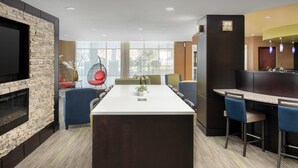 A modern hotel lobby with a central island table, seating area with chairs, a fireplace, and a bar area.